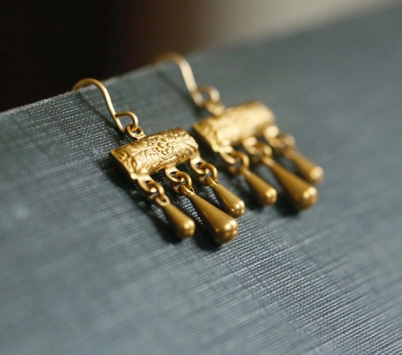May include: A pair of gold-toned drop earrings featuring a rectangular top bar with an embossed floral pattern. Three small, smooth, teardrop-shaped charms hang from each bar, suspended by thin metal loops. The earrings include curved wire hooks.