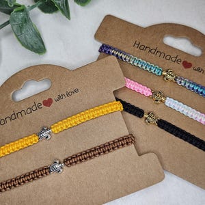 Könnte beinhalten: Fünf geflochtene Armbänder auf zwei braunen Pappkarten mit der Aufschrift Handmade with love. Die Armbänder haben kleine Schildkröten-Anhänger aus gold- und silberfarbenem Metall. Die Bänder sind gelb, braun, schwarz, rosa und mehrfarbig.
