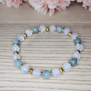Könnte beinhalten: Ein Armband mit hellblauen und weißen opalartigen Perlen, unterbrochen von goldfarbenen Elementen. Das Armband liegt auf einer hellen Holzoberfläche, im Hintergrund rosa Blüten. Die Perlen haben einen Durchmesser von etwa 0,8 cm.
