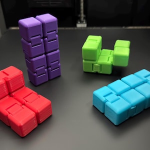 May include: Four colourful fidget toys made of plastic. The toys are in the shapes of a chair, a couch, a tower, and a rectangle. The chair is red, the couch is blue, the tower is purple, and the rectangle is green.