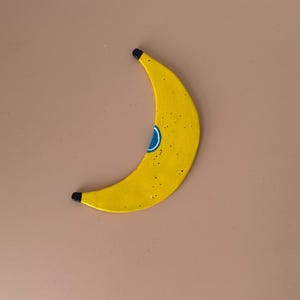 Könnte beinhalten: Ein gebogenes, bananenförmiges Objekt in leuchtendem Gelb mit schwarzen Spitzen an beiden Enden. Die Oberfläche weist kleine schwarze Sprenkel und ein zentrales blaues kreisförmiges Design mit weißem Rand auf. Das Objekt hat eine glatte Oberfläche.