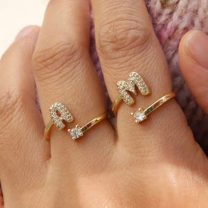 Op de afbeelding: Twee goudkleurige open ringen gedragen aan naast elkaar gelegen vingers. Elke ring heeft een hoofdletter, een A en een M, ingelegd met kleine heldere steentjes. Het andere uiteinde van elke open ring is afgewerkt met een enkele, grotere ronde steen.