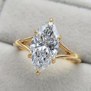Puede incluir: Anillo de compromiso con diamante talla marquesa. El diamante claro está engastado en una banda de oro con un diseño de caña dividida. El anillo se muestra sobre un fondo suave y neutro.