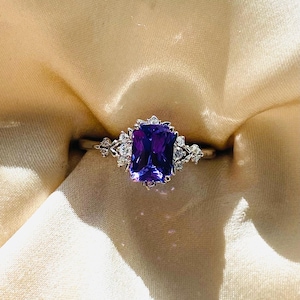 Puede incluir: Un anillo de plata con una gran gema rectangular de color púrpura oscuro, rodeada de piedras más pequeñas y claras. El anillo se muestra sobre una tela suave de color crema, resaltando el brillo y el color de la joya.