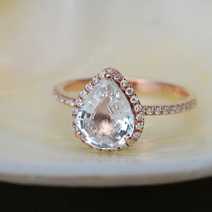 Puede incluir: Un anillo de compromiso de diamante en forma de pera engastado en oro rosa con un halo de diamantes m&aacute;s peque&ntilde;os alrededor de la piedra central.