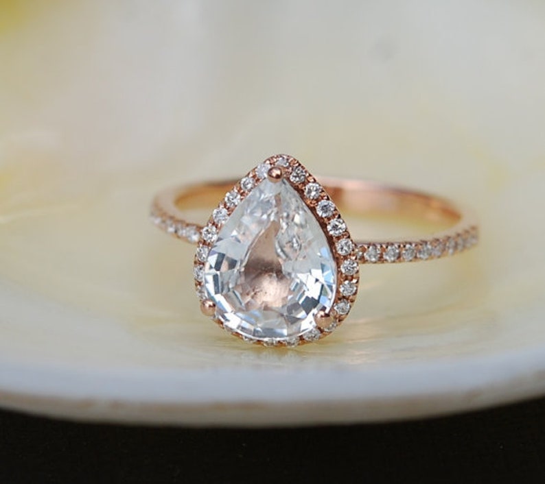 Puede incluir: Un anillo de compromiso de diamante en forma de pera engastado en oro rosa con un halo de diamantes m&aacute;s peque&ntilde;os alrededor de la piedra central.