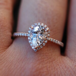 May include: A pear-shaped diamond engagement ring with a halo setting. The ring is set in rose gold and features a band of small diamonds.