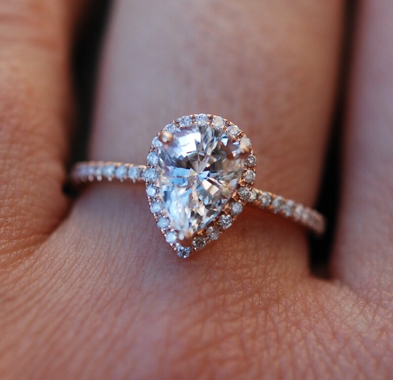 May include: A pear-shaped diamond engagement ring with a halo setting. The ring is set in rose gold and features a band of small diamonds.