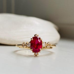 May include: A gold ring featuring a central oval-cut vibrant red gemstone held by prongs. The band is accented with small, clear round-cut stones arranged in clusters on either side of the main setting, creating a detailed and symmetrical design.