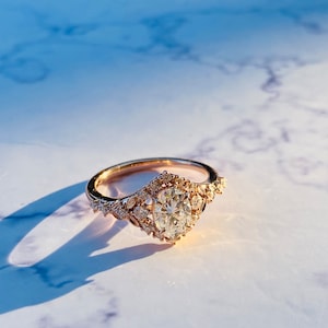 Puede incluir: Un anillo de compromiso de oro rosa con un gran diamante de forma ovalada rodeado de diamantes más pequeños. El anillo tiene un delicado diseño floral.