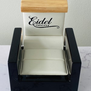 Puede incluir: Caja de anillo negra con interior blanco y tapa de madera. La caja tiene el texto "Eidel Precious" en la parte superior.