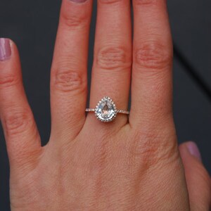 Puede incluir: Un anillo de compromiso de forma de pera con una piedra central transparente rodeada por un halo de piedras m&aacute;s peque&ntilde;as. El anillo est&aacute; engastado en oro rosa y se lleva en una mano con esmalte de u&ntilde;as rosa claro.