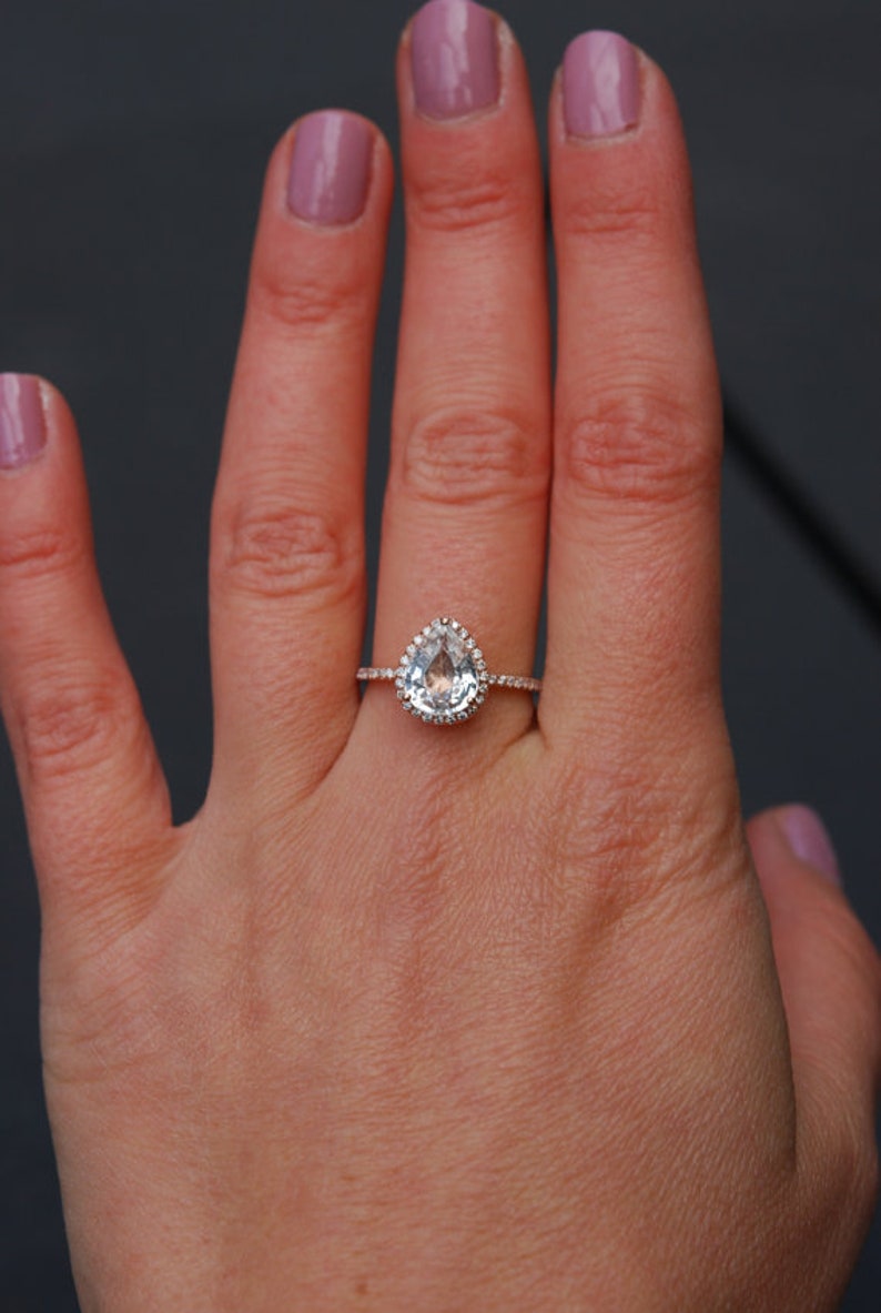 Puede incluir: Un anillo de compromiso de forma de pera con una piedra central transparente rodeada por un halo de piedras m&aacute;s peque&ntilde;as. El anillo est&aacute; engastado en oro rosa y se lleva en una mano con esmalte de u&ntilde;as rosa claro.