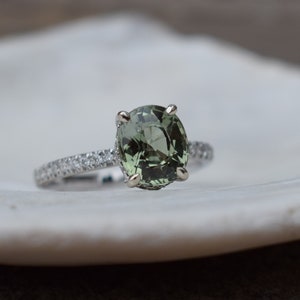 May include: A silver ring with a large, oval-cut, light green gemstone as the centerpiece. The band is encrusted with small, clear gemstones. The ring is set on a white, textured surface, creating a contrast between the jewelry and the background.
