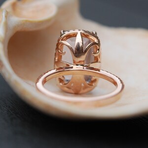 May include: A rose gold engagement ring with a large oval-shaped center stone set in a delicate filigree design. The ring is resting inside a seashell.