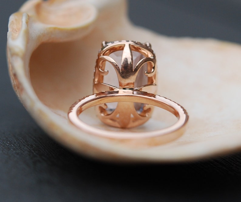 May include: A rose gold engagement ring with a large oval-shaped center stone set in a delicate filigree design. The ring is resting inside a seashell.