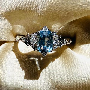 Puede incluir: Un anillo plateado con una gran gema rectangular de color azul claro como pieza central. El anillo está adornado con gemas más pequeñas y transparentes, y detalles decorativos en forma de hojas en los laterales. El anillo se encuentra sobre una tela beige suave.