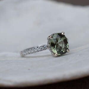 May include: A silver ring with a large, oval-cut, olive-green gemstone as the centerpiece. The band is adorned with small, clear gemstones. The ring is set against a white, textured background, showcasing the jewelry's details.