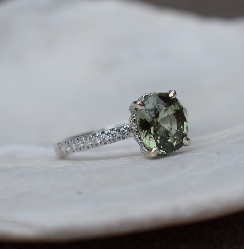 May include: A silver ring with a large, oval-cut, olive-green gemstone as the centerpiece. The band is adorned with small, clear gemstones. The ring is set against a white, textured background, showcasing the jewelry's details.