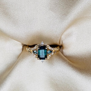 May include: A gold ring with a large emerald green gemstone surrounded by smaller diamonds. The ring has a delicate, intricate design.