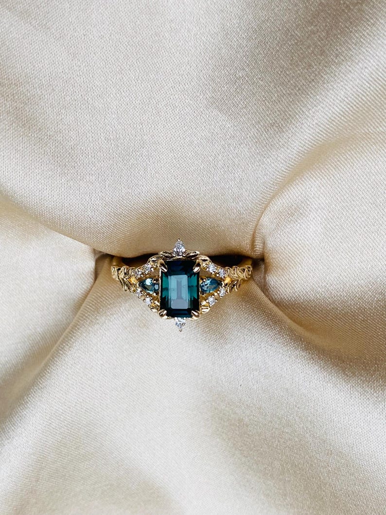 May include: A gold ring with a large emerald green gemstone surrounded by smaller diamonds. The ring has a delicate, intricate design.