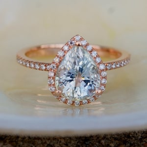 May include: A pear-shaped diamond engagement ring with a halo setting in rose gold. The ring is set with smaller diamonds around the center stone.