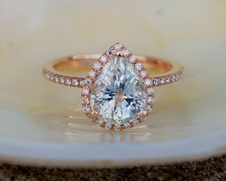 May include: A pear-shaped diamond engagement ring with a halo setting in rose gold. The ring is set with smaller diamonds around the center stone.