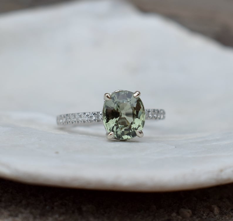 May include: A silver ring with a large, oval-cut, light green gemstone as the centerpiece. The band is encrusted with small, clear gemstones. The ring is set against a blurred, off-white background, highlighting the jewelry's details.