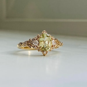 Puede incluir: Un anillo de oro con una gran piedra preciosa ovalada verde rodeada de diamantes más pequeños. El anillo tiene un delicado diseño floral.