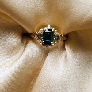 May include: A gold ring with a large emerald green gemstone surrounded by smaller diamonds and blue gemstones. The ring is set on a white satin background.