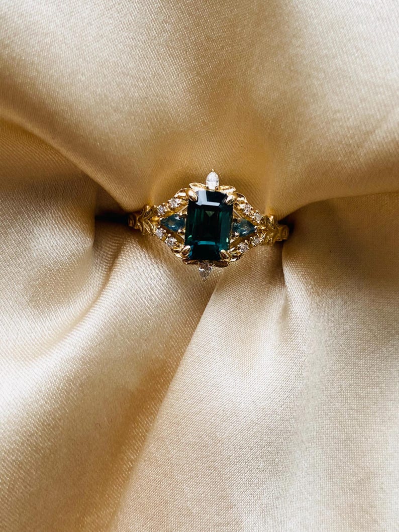 May include: A gold ring with a large emerald green gemstone surrounded by smaller diamonds and blue gemstones. The ring is set on a white satin background.