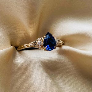 Puede incluir: Un anillo de oro con una piedra preciosa de zafiro azul en forma de pera rodeada de pequeñas piedras preciosas transparentes.