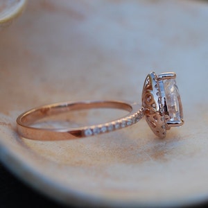 May include: A rose gold engagement ring with a large, clear, oval-shaped center stone surrounded by a halo of smaller diamonds. The band is also set with smaller diamonds.