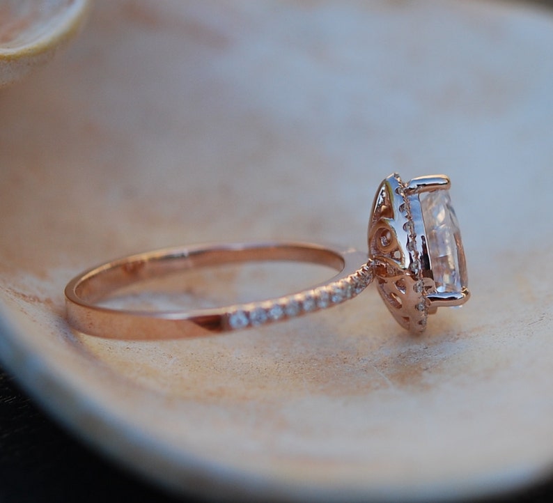 May include: A rose gold engagement ring with a large, clear, oval-shaped center stone surrounded by a halo of smaller diamonds. The band is also set with smaller diamonds.