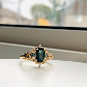 May include: A gold ring with a large emerald green gemstone surrounded by smaller diamonds and blue gemstones. The ring has a delicate floral design.