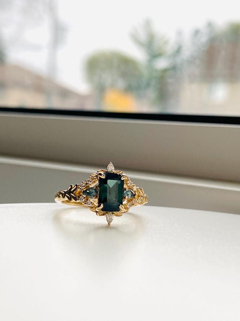 May include: A gold ring with a large emerald green gemstone surrounded by smaller diamonds and blue gemstones. The ring has a delicate floral design.