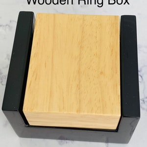May include: A wooden ring box with a black exterior and a light wood interior. The box is closed and the lid is visible.