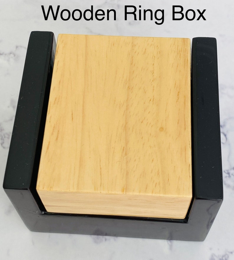 May include: A wooden ring box with a black exterior and a light wood interior. The box is closed and the lid is visible.
