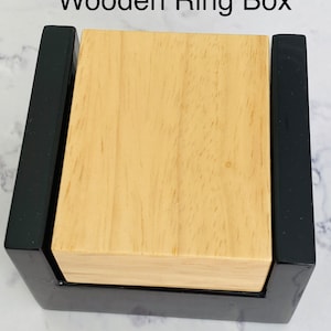 Puede incluir: Una caja de madera para anillos con un exterior negro y un interior de madera clara. La caja est&aacute; cerrada y la tapa es visible.