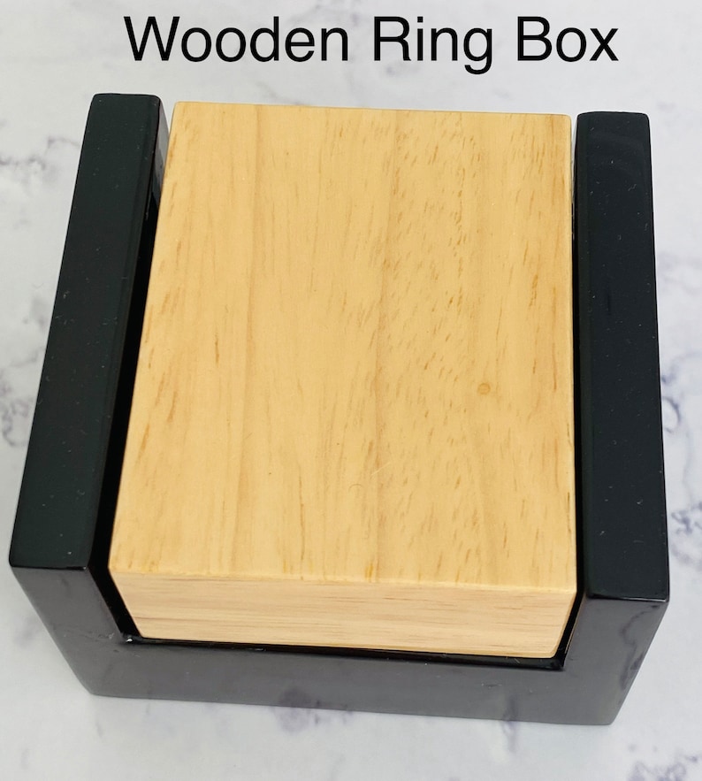 Puede incluir: Una caja de madera para anillos con un exterior negro y un interior de madera clara. La caja est&aacute; cerrada y la tapa es visible.