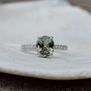 May include: A silver ring with a large, oval-cut, olive-green gemstone as the focal point. The band is adorned with small, clear gemstones. The ring is set against a white background, highlighting the jewelry's details.