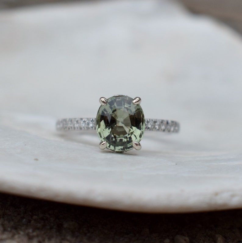 May include: A silver ring with a large, oval-cut, olive-green gemstone as the focal point. The band is adorned with small, clear gemstones. The ring is set against a white background, highlighting the jewelry's details.