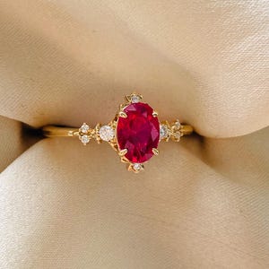May include: A gold ring featuring an oval-cut, deep red gemstone as the centerpiece. The ring is adorned with smaller, clear gemstones and delicate gold detailing on the band, creating an elegant and classic design. The ring is set against a neutral background.