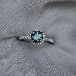 May include: A silver ring featuring a round, teal blue gemstone encircled by a halo of smaller diamonds.