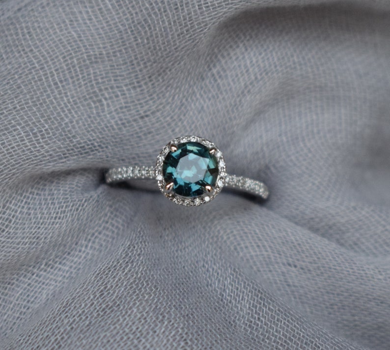 May include: A silver ring featuring a round, teal blue gemstone encircled by a halo of smaller diamonds.