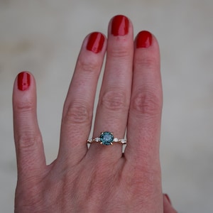 May include: A gold ring with a teal gemstone surrounded by smaller diamonds. The ring is on a person's finger.