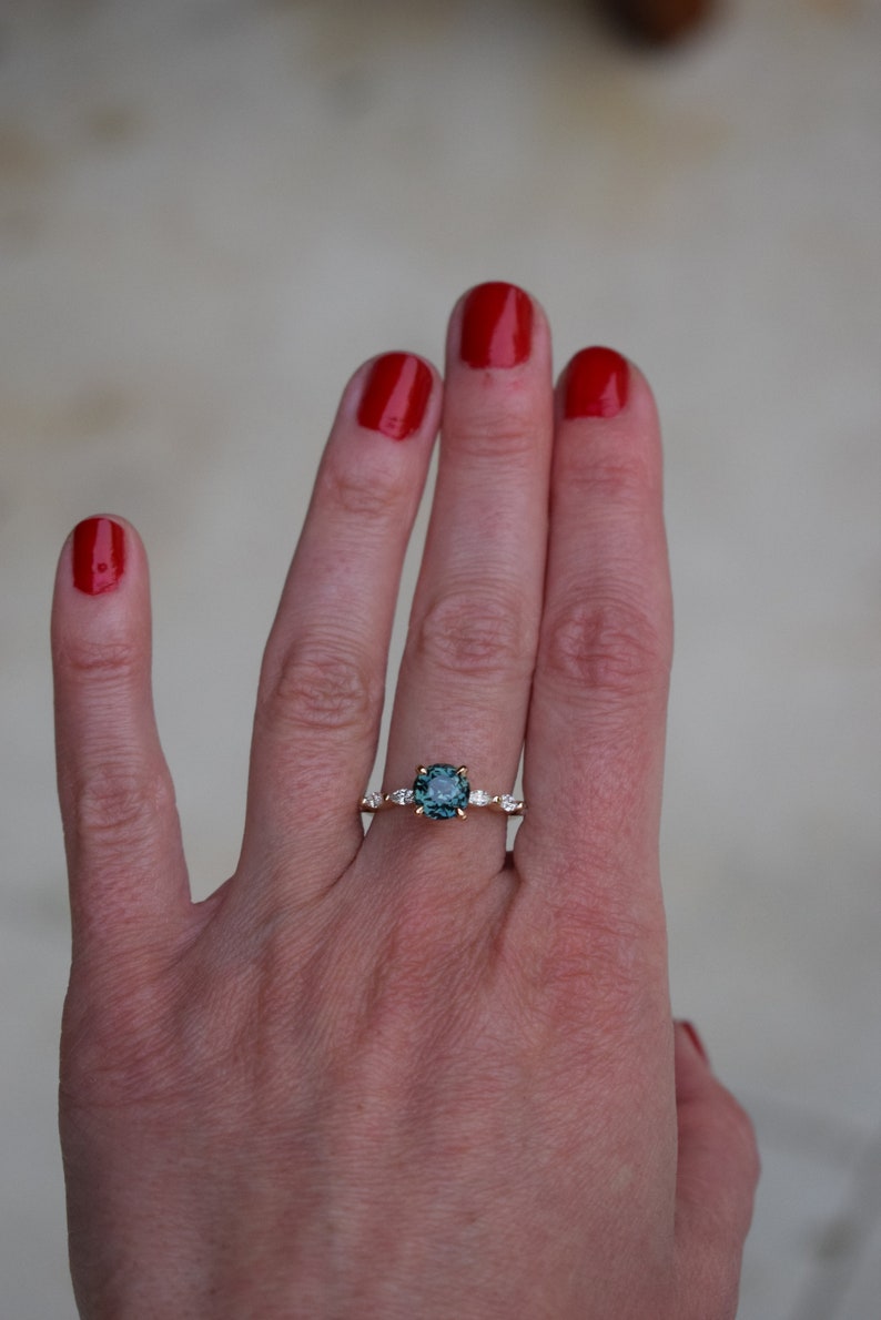 May include: A gold ring with a teal gemstone surrounded by smaller diamonds. The ring is on a person's finger.