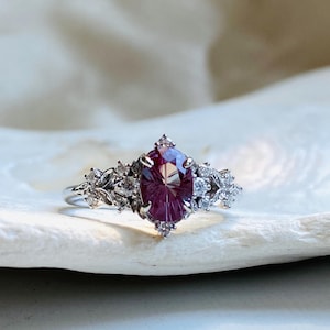 Puede incluir: Un anillo de plata con una gran piedra preciosa ovalada de color púrpura oscuro. El anillo está adornado con piedras preciosas claras más pequeñas y delicados diseños florales o de hojas en la banda, creando una apariencia elegante y detallada.