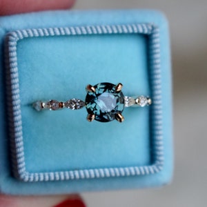 May include: A teal blue gemstone ring with a gold band and a row of small diamonds. The ring is in a light blue ring box.