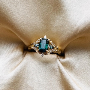 May include: A gold ring with a large emerald green gemstone surrounded by smaller diamonds. The ring is set on a white satin background.